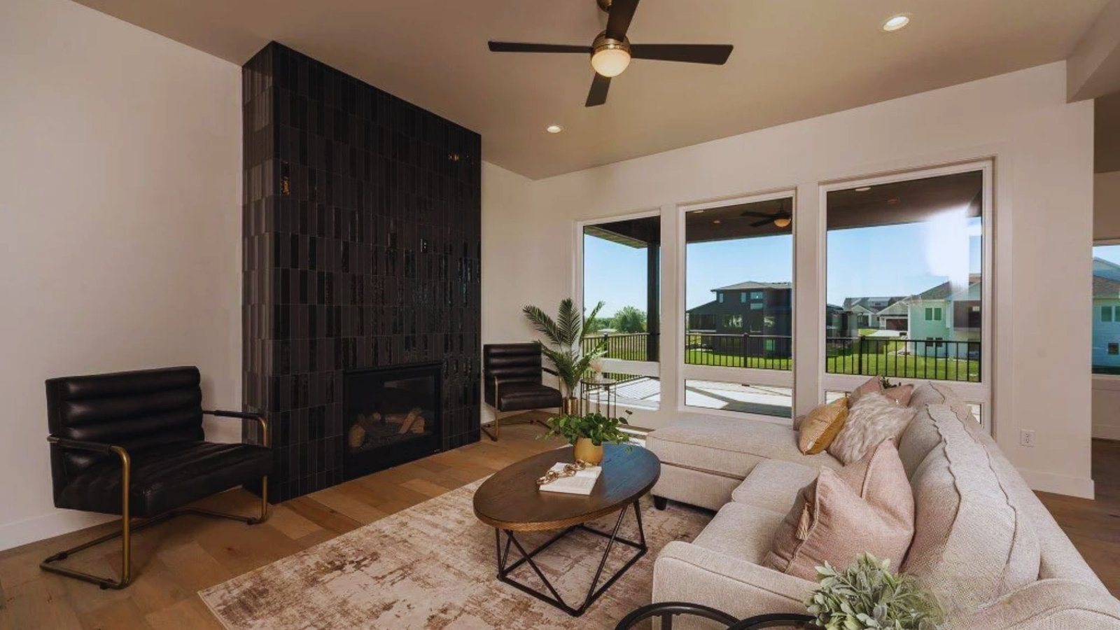 A modern living room with a large beige sectional sofa, dark accent chairs, and a black tiled fireplace. Bright natural light streams through three tall windows, revealing a view of a patio and lawn. A ceiling fan adds a cozy touch, while a coffee table with plants and books completes the inviting setup.