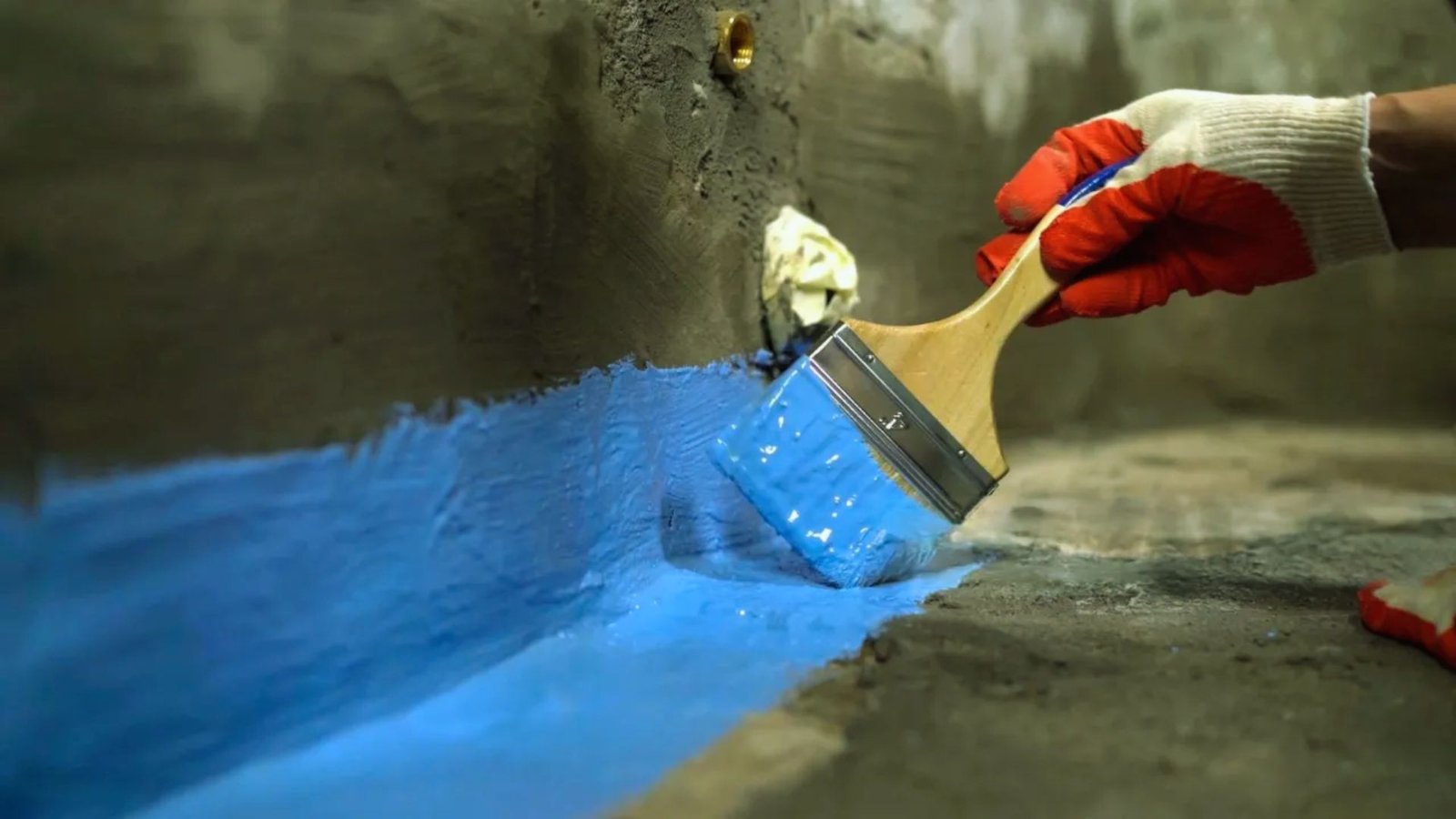 A hand wearing red and white gloves uses a brush to apply bright blue paint on a gray concrete surface, suggesting a renovation or artistic project.