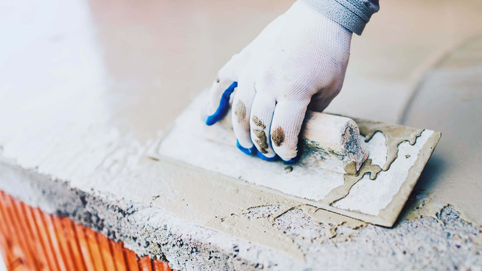 A gloved hand smooths wet cement on a surface using a trowel, showcasing construction work. The focus is on the texture of the cement and the precision involved.