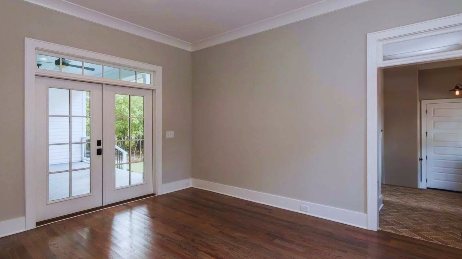 Spacious room with light gray walls, wooden floor, and French doors opening to a porch. The space feels bright and inviting with natural sunlight.