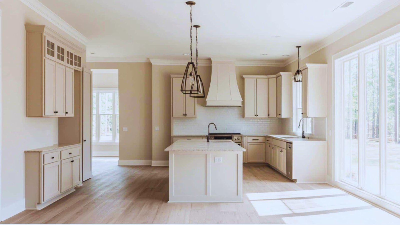Spacious, sunlit kitchen with cream cabinets, island with pendant lights, wood floors, and large windows. Simple, modern, and inviting design.